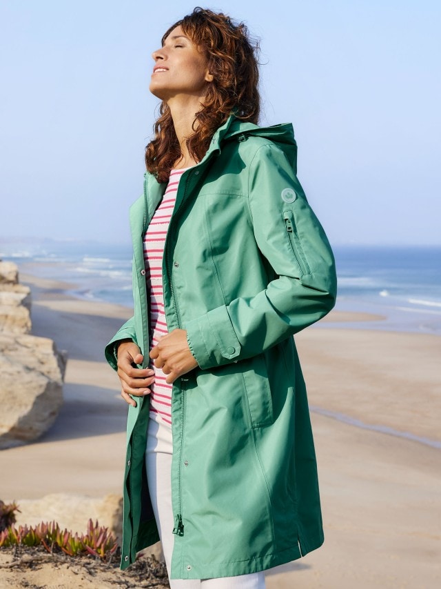 Frau am Strand trägt eine grüne Jacke, genießt Sonne, Hintergrund aus Meer und Felsen.