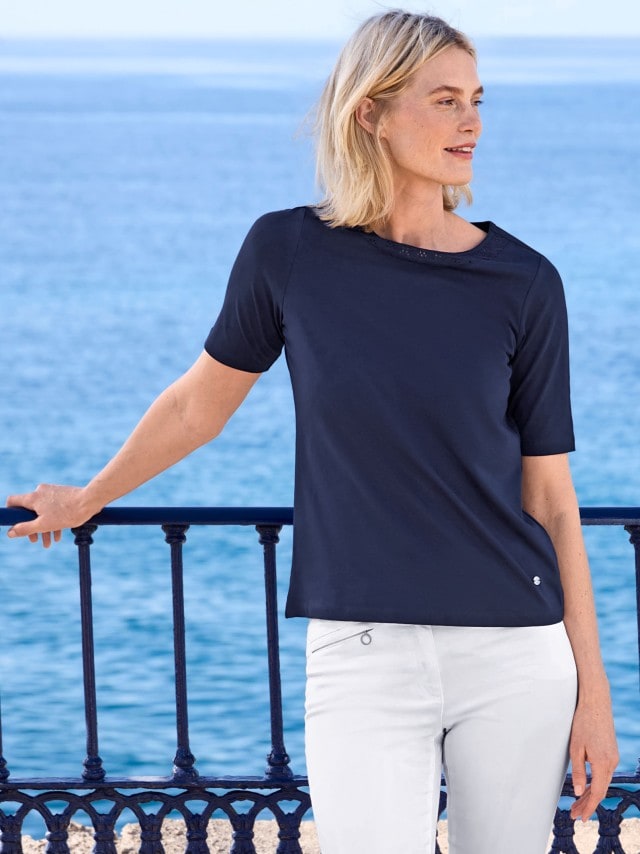 Frau in einem Marineblauem T-Shirt und weißer Hose hält sich an einem Geländer an der Küste fest