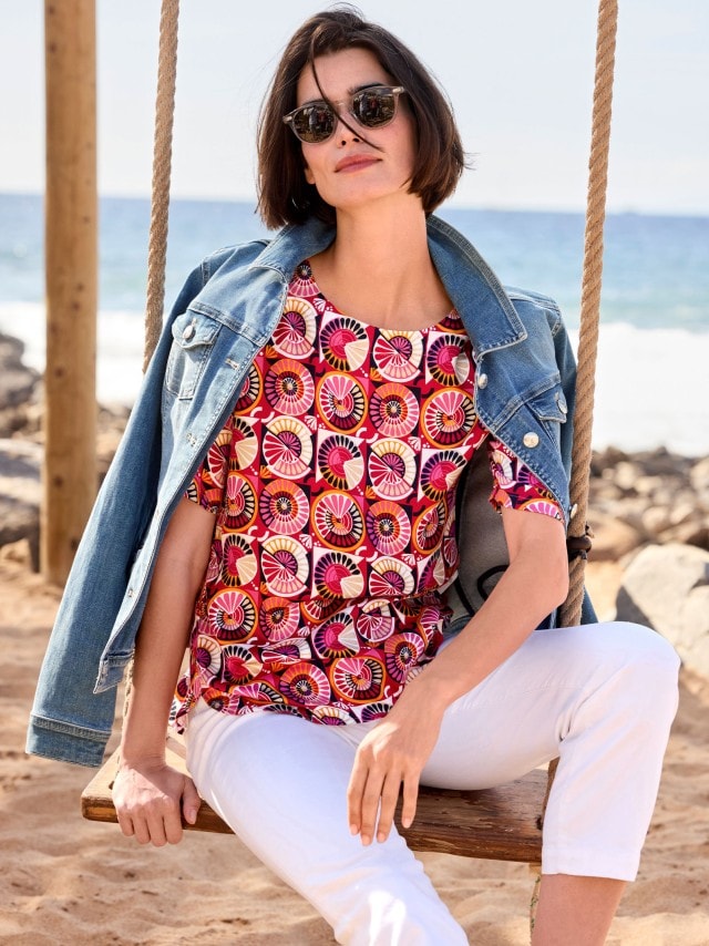 Frau mit Sonnenbrille sitzt auf einer Schaukel am Strand, trägt eine rotgemusterte T-Shirt-Bluse, weiße Hose und hat eine Jeansjacke über ihren Schultern hängen