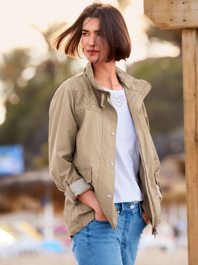 Frau steht am Strand und trägt eine Baumwolljacke Ajour in Taupe, darunter ein weißes Shirt und steckt ihre Hände in die Taschen einer blauen Jeans