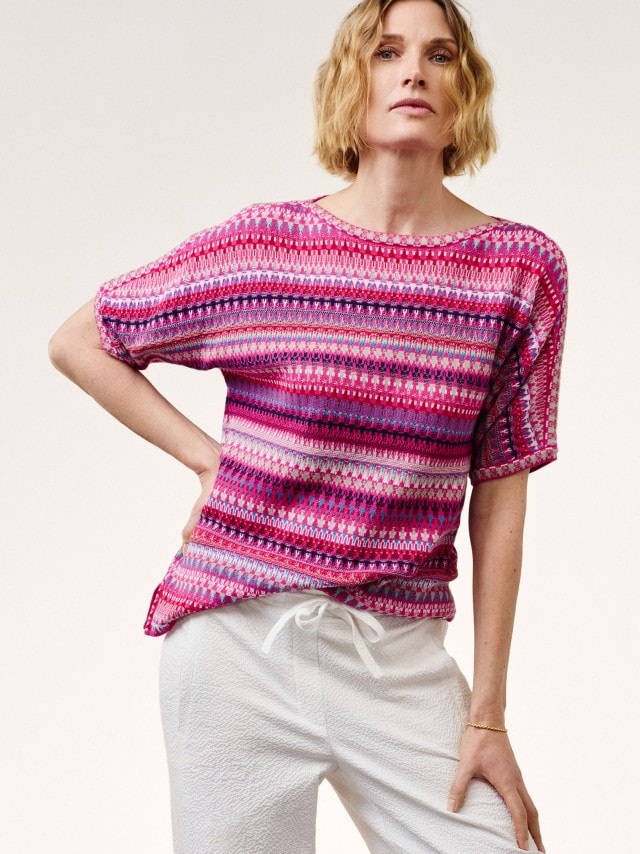 Blonde Frau vor neutralem Hintergrund trägt den 
Kimono Pullover Baumwolljacquard in Fuchsia dazu eine weiße Hose
