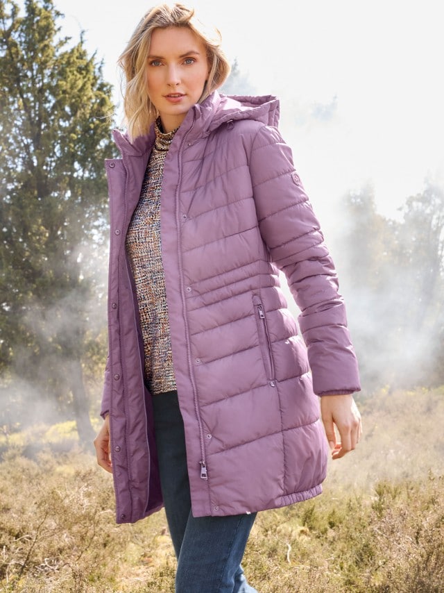 Eine Person trägt eine lilafarbene, gesteppte Winterjacke mit Kapuze, kombiniert mit Jeans und einem gemusterten Pullover. Der Stil wirkt modern und gemütlich, passend für kühle Tage im Freien.