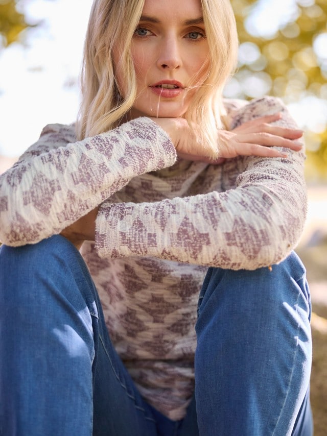 Getragen wird ein langärmliges Oberteil mit lila-weißem Muster und eine Jeans. Der Stil wirkt entspannt und angenehm lässig für den Alltag.