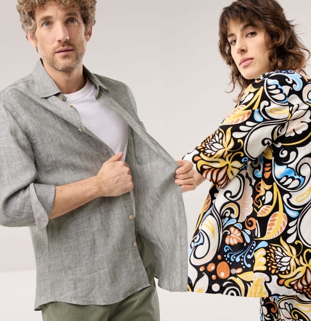 Ein Mann trägt ein offenes Leinenhemd in der Farbe Schilf, eine Frau in einer gemusterten Hemdbluse aus Leinenmix.