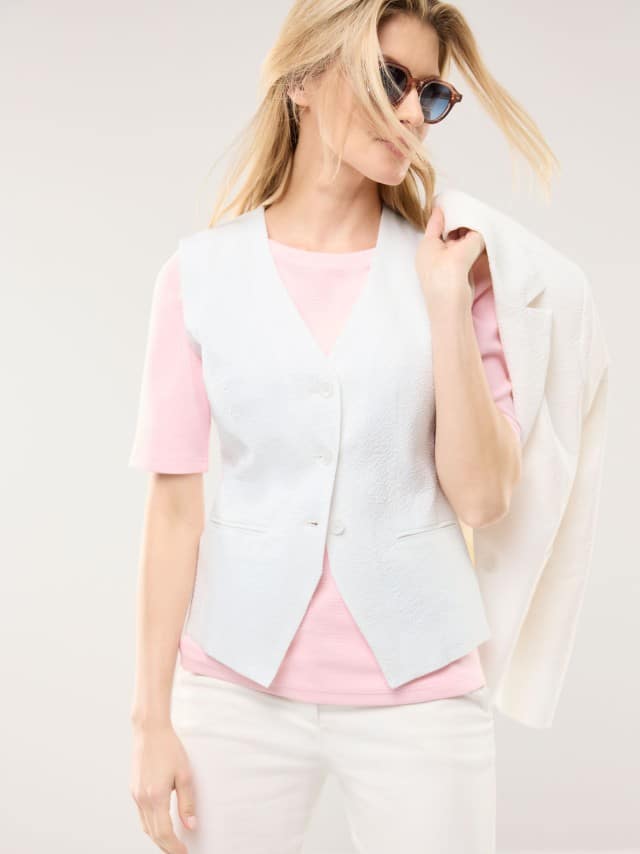Eine Frau trägt ein helles, frisches Outfit aus weißer Weste, weißem Blazer und rosa Oberteil, das einen modernen und entspannt‑lebendigen Eindruck vermittelt.