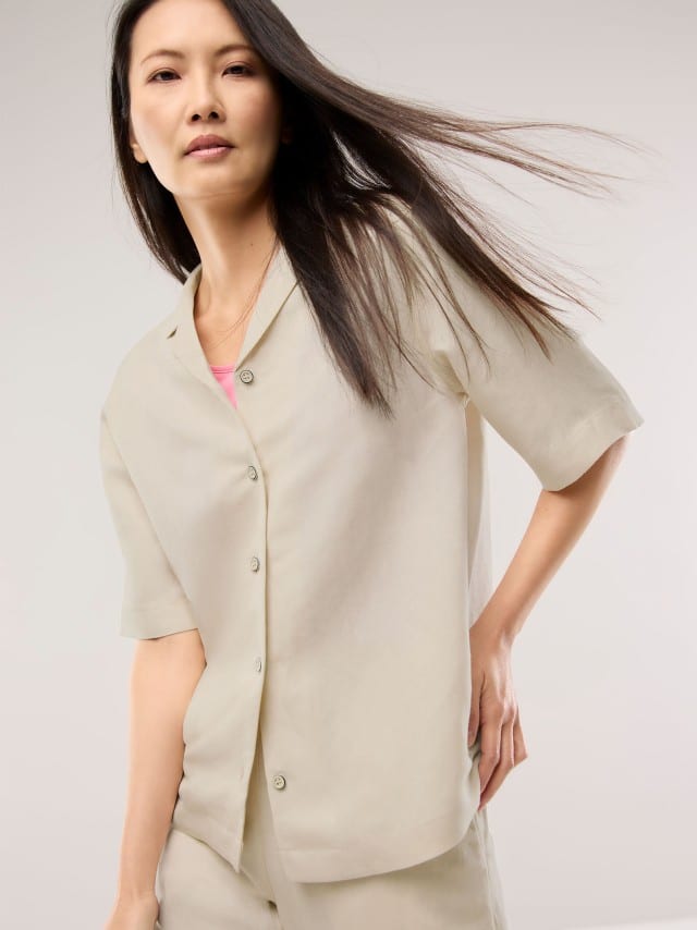 Eine Frau trägt ein helles, locker fallendes Set aus Kurzarmhemd und passender Hose über einem rosa Oberteil, was einen minimalistischen, weichen und entspannt‑leichten Eindruck vermittelt.