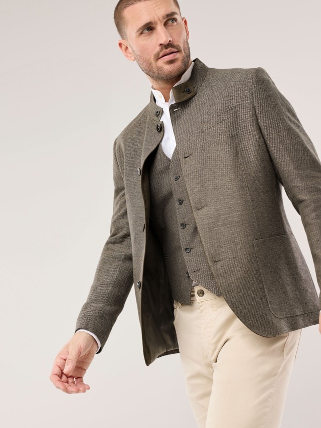 Ein Mann trägt eine braune Strukturjacke mit passender Weste zu einem weißen Hemd und hellen Hosen, was traditionell‑modern wirkt und eine ruhige, stilvolle Stimmung vermittelt.