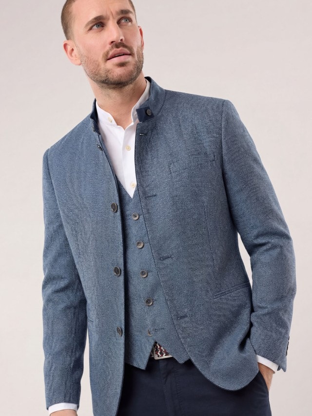 Ein Mann trägt eine blau strukturierte Jacke mit passender Weste zu einem weißen Hemd, was modern‑traditionell wirkt und eine ruhige, gepflegte Stimmung vermittelt.