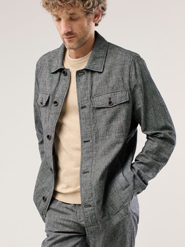 Ein Mann trägt ein grau kariertes Set aus Jacke und Hose über einem hellen Pullover, was modern, dezent und entspannt wirkt.