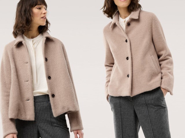 Eine Frau trägt einen beigen Kurzblazer mit Knöpfen über einer weißen Bluse. Dazu kombiniert sie eine dunkelgraue Bundfaltenhose mit feinen Nadelstreifen für einen eleganten Business-Look.