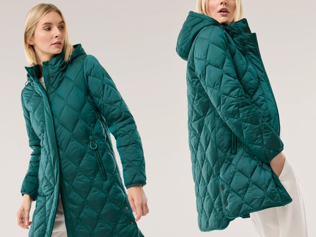Eine Frau trägt eine lange, smaragdgrüne Steppjacke mit Kapuze und Reißverschluss. Der Look ist warm, modern und sportlich-elegant.