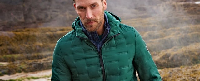 Mann steht mit einer grünen Outdoorjacke bekleidet in einer nebligen Landschaft vor mosigen Felsen