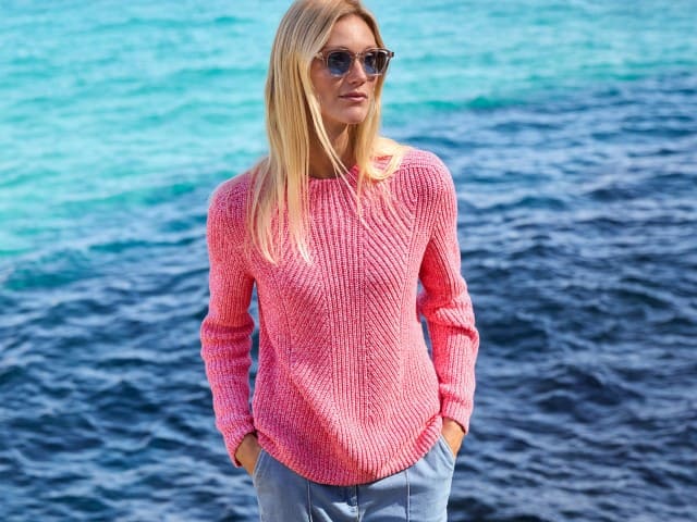 Eine blonde Frau mit Sonnenbrille steht am Wasser und trägt einen Pullover in Rosa, dazu eine Jeans.