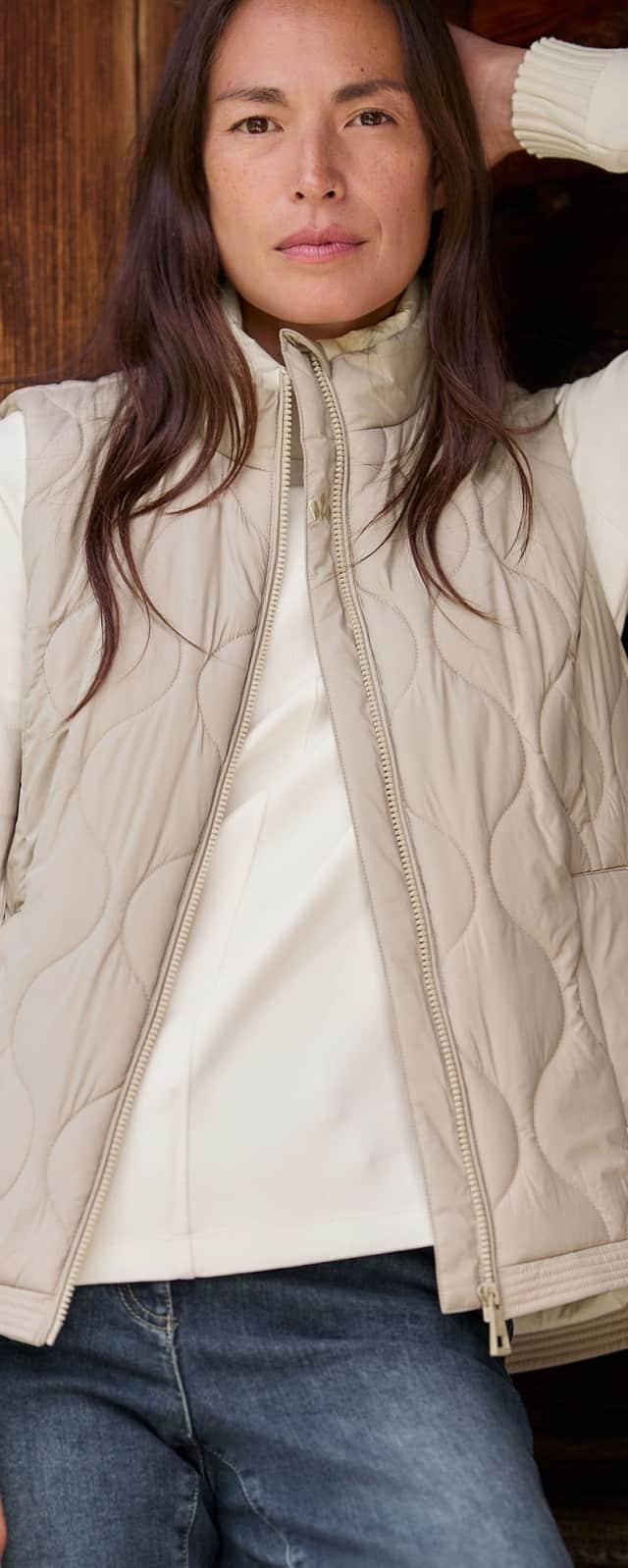 Eine Frau steht vor einer Holzwand und trägt die Wellensteppweste in Toffee/Creme, darunter ein weißes Sweatshirt und dazu eine Jeans.