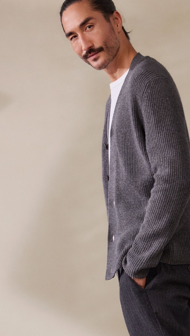 Ein Mann trägt ein weißes T-Shirt mit einer grob gestrickten, grauen Strickjacke. Dazu kombiniert er eine dunkle Hose für einen entspannten, modernen Look.