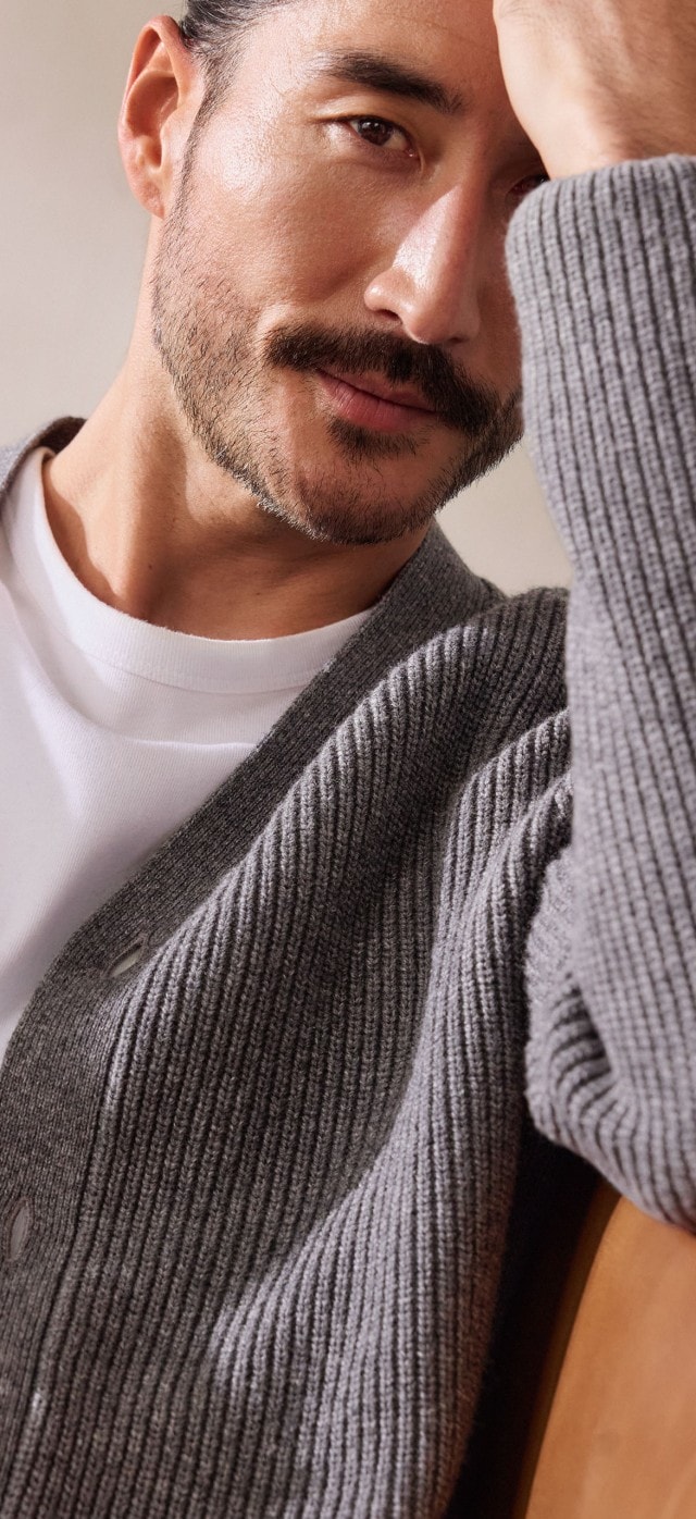 Ein Mann trägt ein weißes T-Shirt mit einer grob gestrickten, grauen Strickjacke. Dazu kombiniert er eine dunkle Hose für einen entspannten, modernen Look.