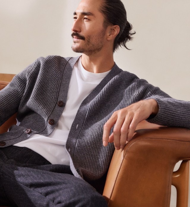Ein Mann trägt ein weißes T-Shirt mit einer grob gestrickten, grauen Strickjacke. Dazu kombiniert er eine dunkle Hose für einen entspannten, modernen Look.