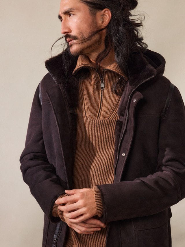 Ein Mann trägt einen braunen Strickpullover mit Reißverschlusskragen unter einer dunkelbraunen Jacke mit Fellfutter. Der Look wirkt warm, robust und zugleich stilvoll.