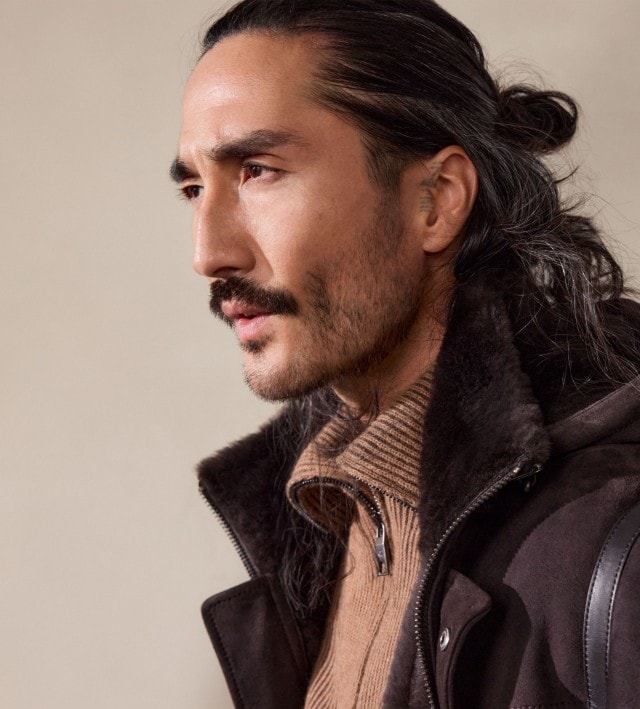 Ein Mann trägt einen braunen Strickpullover mit Reißverschlusskragen unter einer dunkelbraunen Jacke mit Fellfutter. Der Look wirkt warm, robust und zugleich stilvoll.