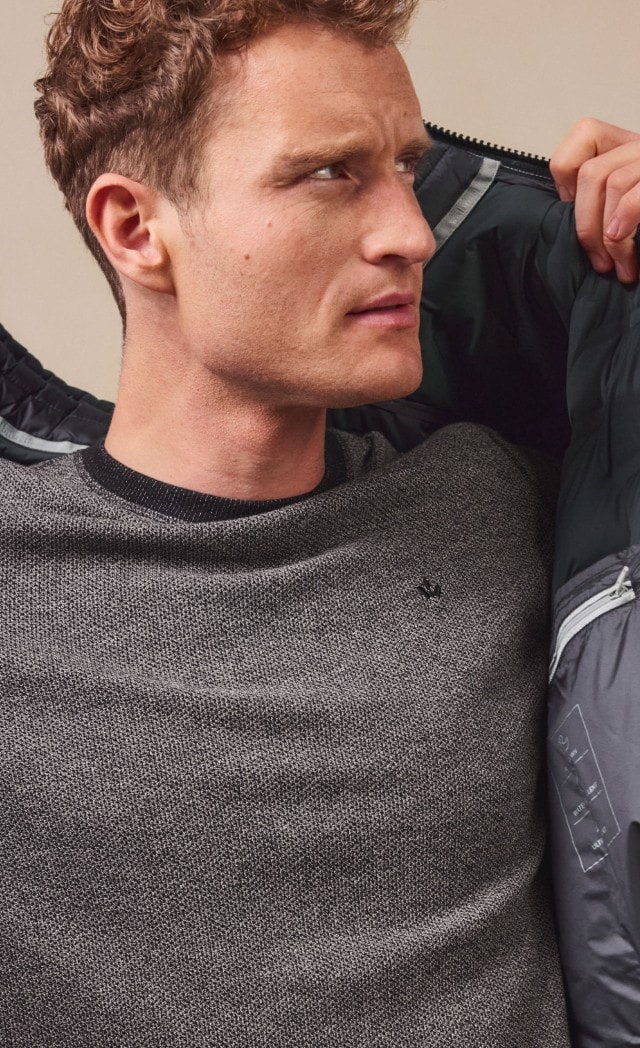 Ein Mann trägt einen grauen Strickpullover, kombiniert mit einer hellgrauen Steppjacke. Der Look wirkt modern, schlicht und zugleich sportlich-elegant.