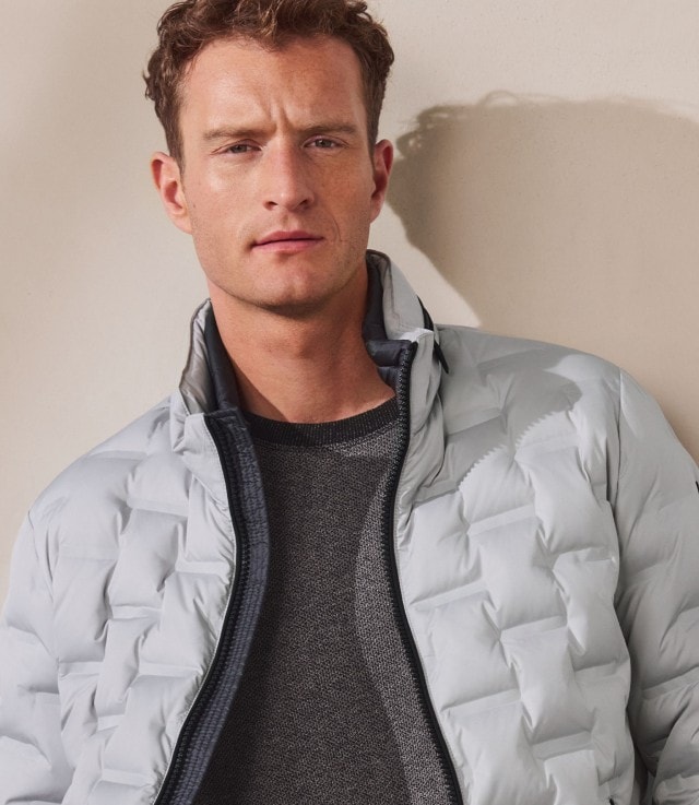 Ein Mann trägt einen grauen Strickpullover, kombiniert mit einer hellgrauen Steppjacke. Der Look wirkt modern, schlicht und zugleich sportlich-elegant.