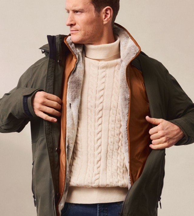 Ein Mann trägt einen dicken cremefarbenen Zopfmuster-Pullover unter einer olivgrünen Outdoor-Jacke mit warmem Innenfutter. Das Outfit ist winterlich, sportlich und funktional.