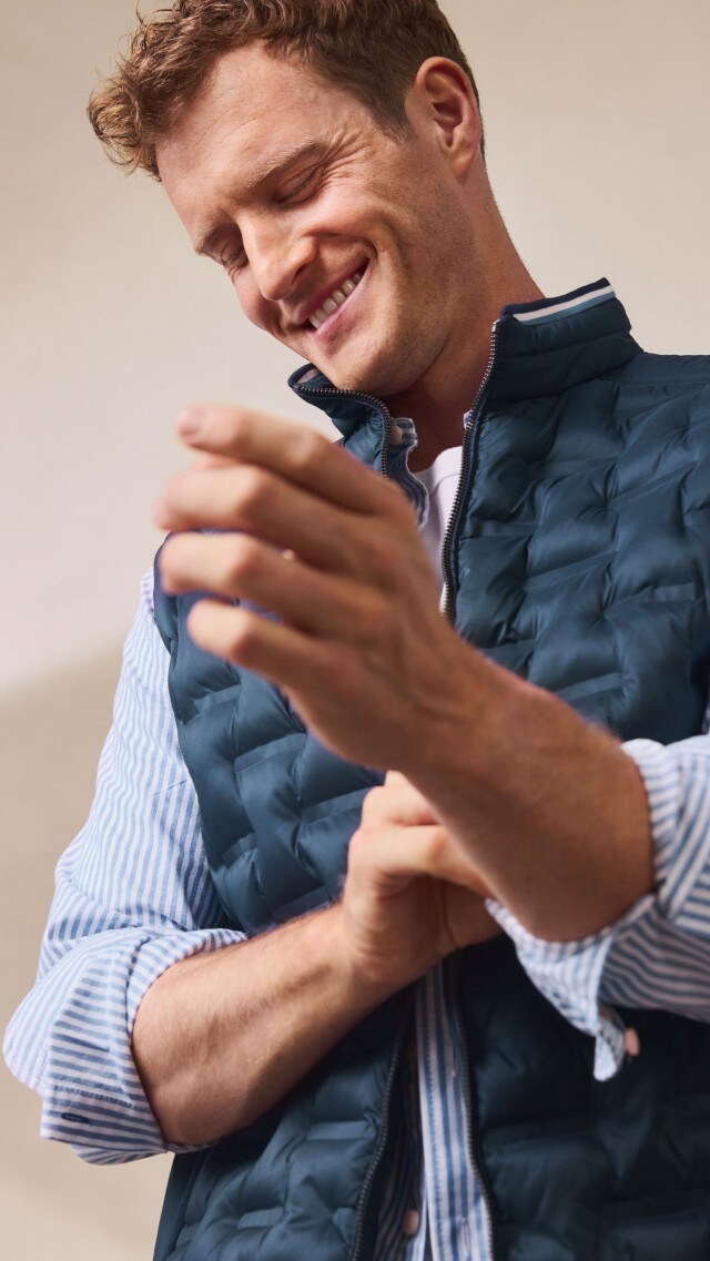 Ein Mann trägt eine gestreifte Hemdbluse mit weißem T-Shirt darunter und eine dunkelblaue Steppweste. Dazu kombiniert er eine beige Hose für ein sportlich-schickes Outfit.