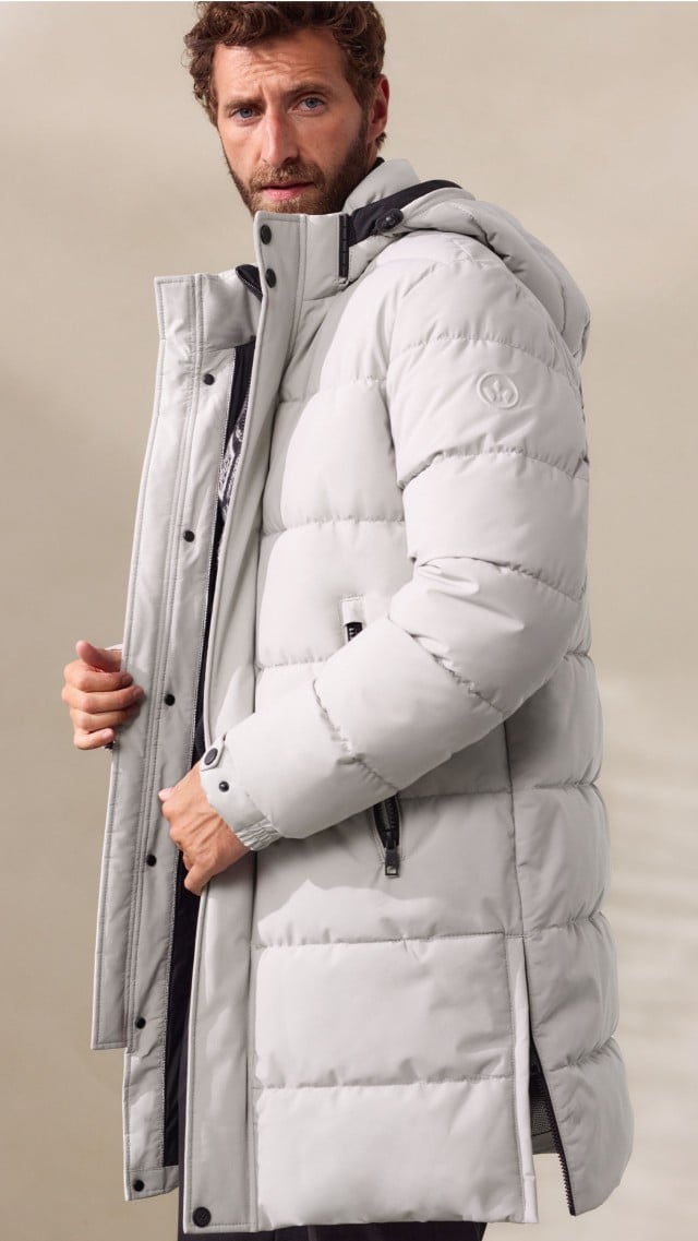 Ein Mann trägt einen schwarzen Pullover mit grafischem Muster unter einem langen, hellgrauen Steppmantel. Der Look ist winterlich, modern und zugleich sportlich-elegant.