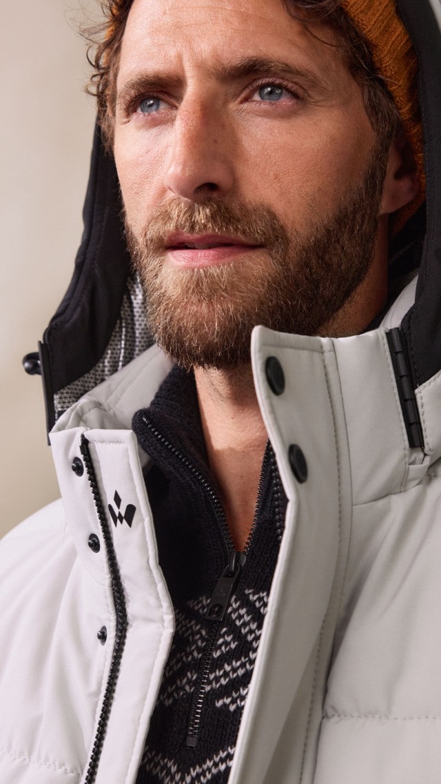 Ein Mann trägt einen schwarzen Pullover mit grafischem Muster unter einem langen, hellgrauen Steppmantel. Der Look ist winterlich, modern und zugleich sportlich-elegant.
