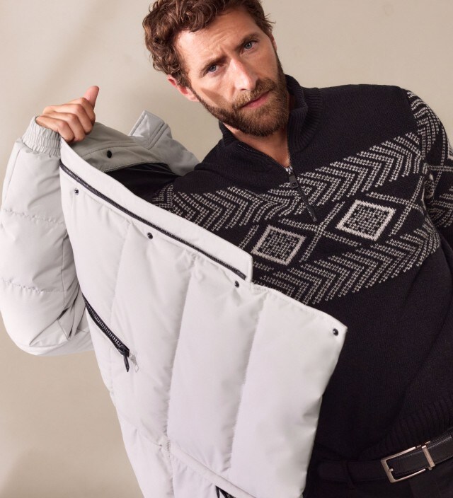 Ein Mann trägt einen schwarzen Pullover mit grafischem Muster unter einem langen, hellgrauen Steppmantel. Der Look ist winterlich, modern und zugleich sportlich-elegant.