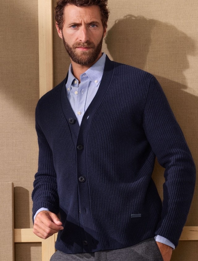 Ein Mann trägt ein hellblaues Hemd unter einer dunkelblauen Strickjacke. Dazu kombiniert er eine graue Hose für einen eleganten, smart-casual Look.