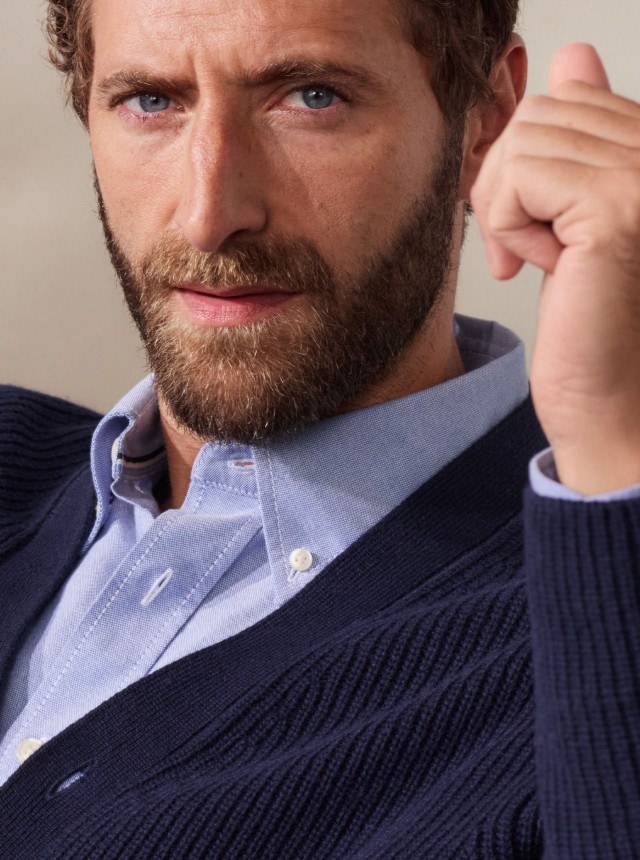 Ein Mann trägt ein hellblaues Hemd unter einer dunkelblauen Strickjacke. Dazu kombiniert er eine graue Hose für einen eleganten, smart-casual Look.
