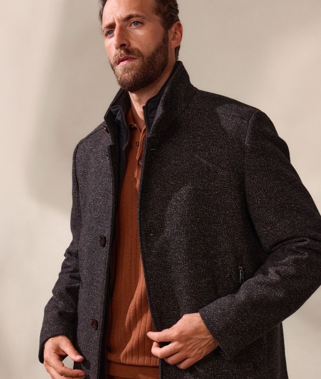 Ein Mann trägt einen dunkel gemusterten Mantel über einem rostfarbenen Pullover. Der Look wirkt stilvoll, herbstlich und klassisch-elegant.