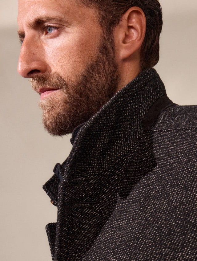 Ein Mann trägt einen dunkel gemusterten Mantel über einem rostfarbenen Pullover. Der Look wirkt stilvoll, herbstlich und klassisch-elegant.