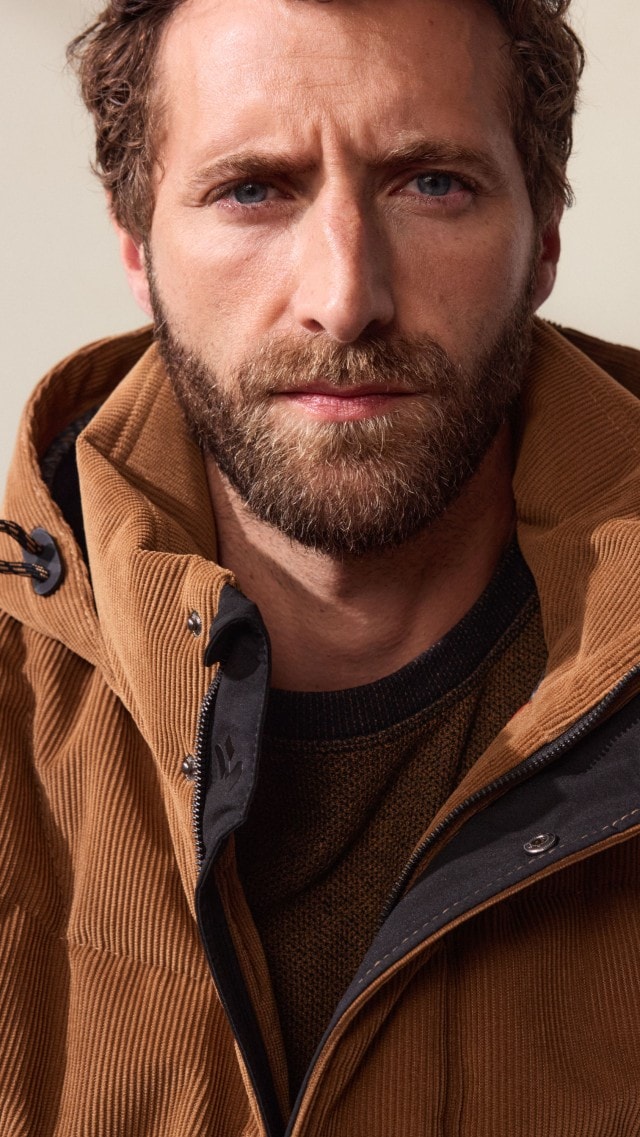 Ein Mann trägt einen braunen Pullover, kombiniert mit einer passenden wattierten Cordjacke. Der Look wirkt herbstlich, lässig und trotzdem stilvoll.