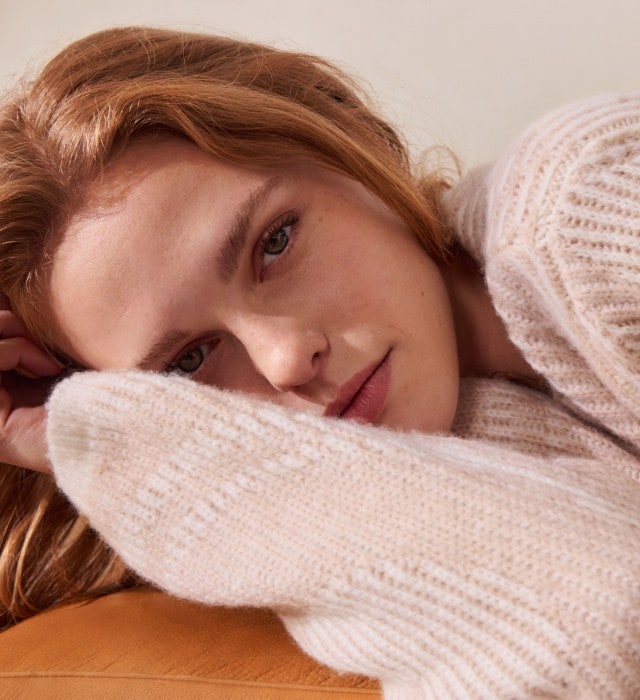 Eine Frau trägt einen weichen, cremefarbenen Strickpullover mit dezentem Muster. Dazu kombiniert sie eine helle Hose für ein gemütliches und elegantes Outfit.