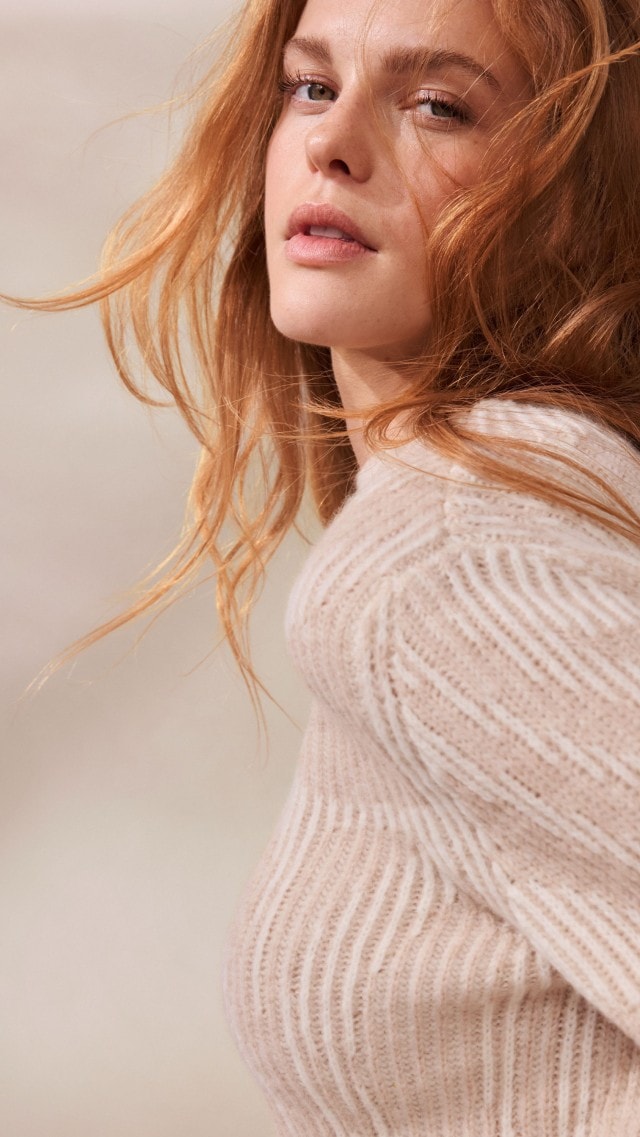 Eine Frau trägt einen weichen, cremefarbenen Strickpullover mit dezentem Muster. Dazu kombiniert sie eine helle Hose für ein gemütliches und elegantes Outfit.