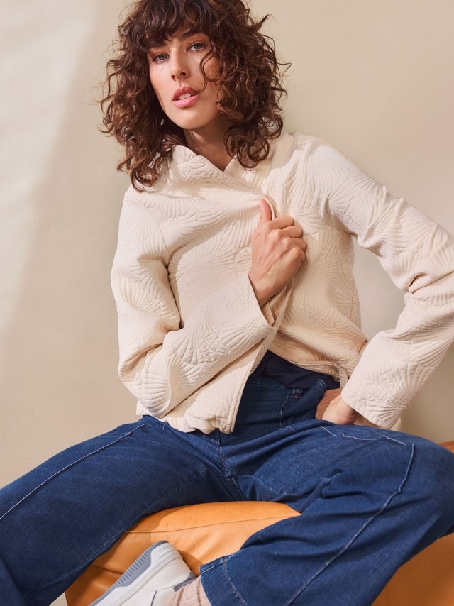 Eine Frau trägt eine helle, strukturierte Jacke mit Reißverschluss. Dazu kombiniert sie eine blaue Jeans und weiße Sneakers für einen modernen, lässigen Look.