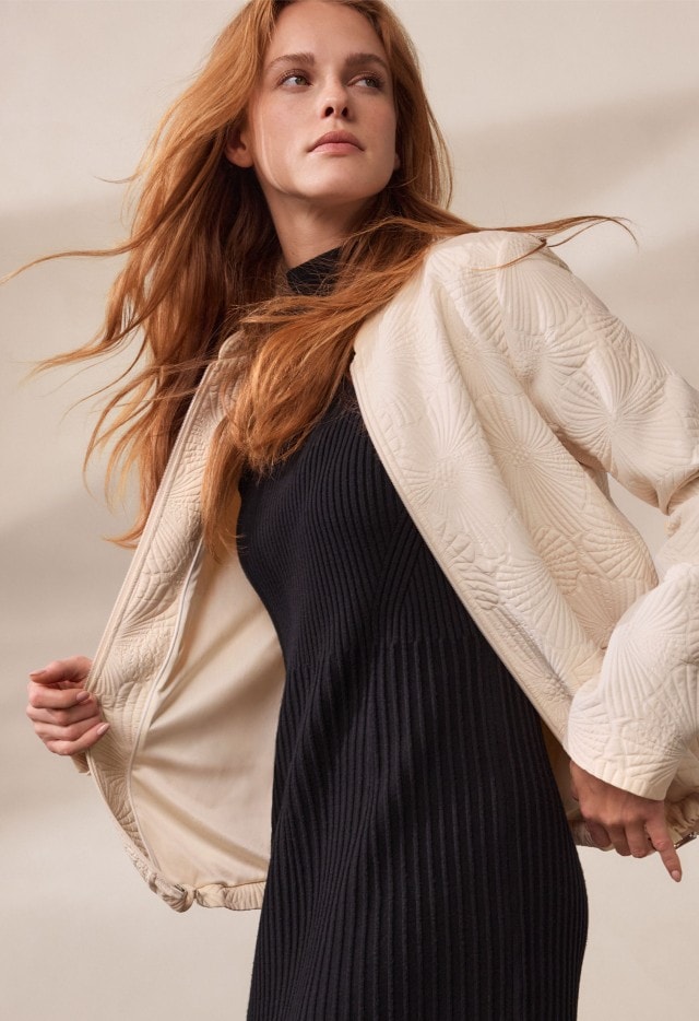 Eine Frau trägt ein elegantes, schwarzes Strickkleid mit langen Ärmeln. Sie kombiniert den Look mit einer hellen Jacke oder warmen Stricksocken für einen gemütlich-modernen Stil.