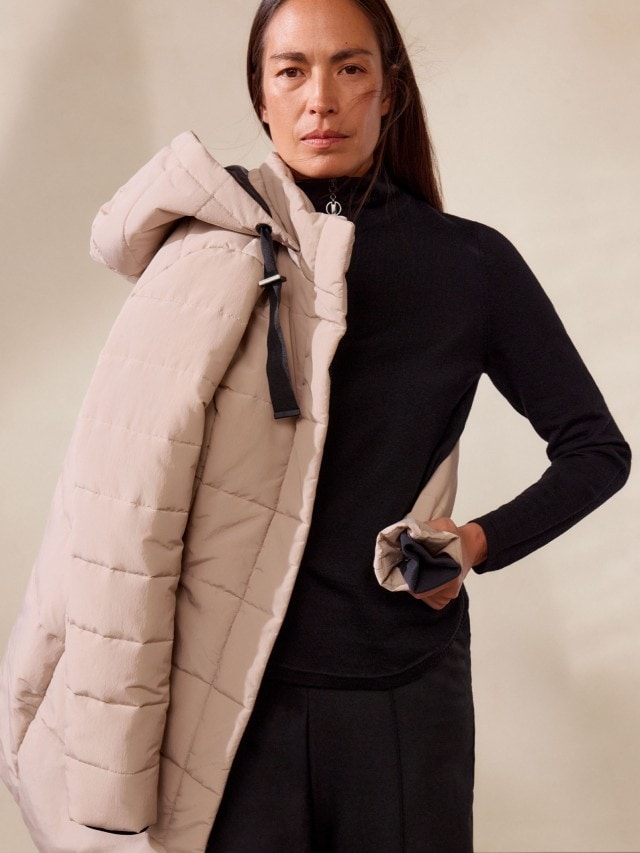 Eine Frau trägt einen beigen Steppmantel mit Kapuze über einem schlichten schwarzen Oberteil. Der Look ist modern, minimalistisch und funktional für kalte Tage.