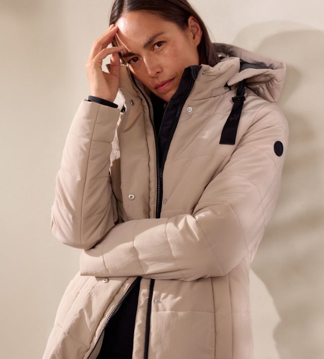 Eine Frau trägt einen beigen Steppmantel mit Kapuze über einem schlichten schwarzen Oberteil. Der Look ist modern, minimalistisch und funktional für kalte Tage.