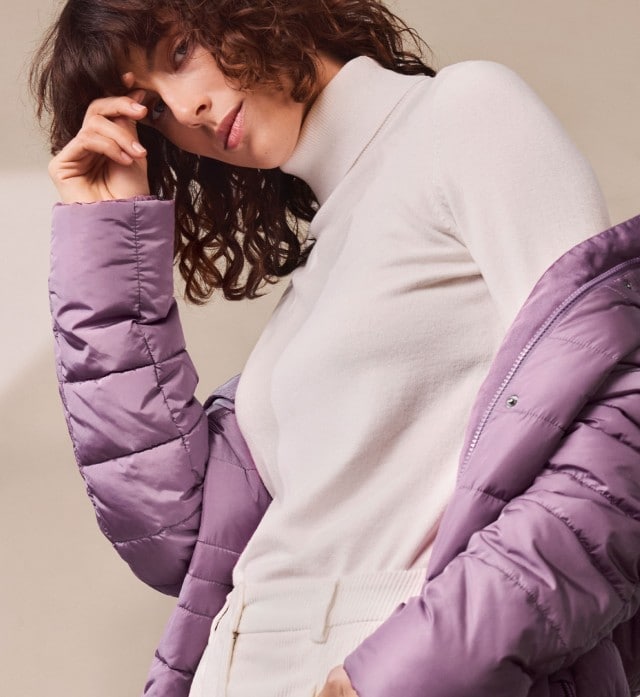Eine Frau trägt einen weißen Rollkragenpullover mit passender heller Hose. Der Look ist elegant, minimalistisch und mit einer lila Steppjacke modern akzentuiert.