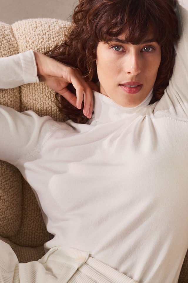 Eine Frau trägt einen weißen Rollkragenpullover mit passender heller Hose. Der Look ist elegant, minimalistisch und mit einer lila Steppjacke modern akzentuiert.