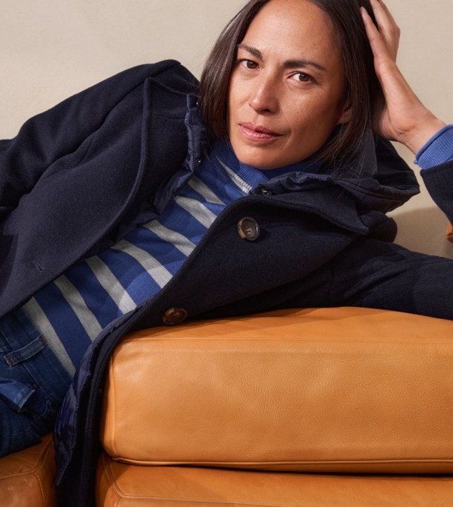 Eine Frau trägt ein blau-weiß gestreiftes Oberteil mit Rollkragen, kombiniert mit Jeans und einem dunkelblauen Mantel. Der Look ist leger, maritim inspiriert und modern.