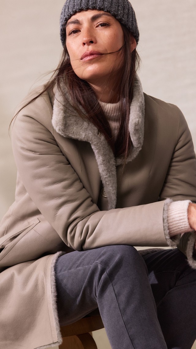 Eine Frau trägt einen hellgrauen Mantel mit weichem Fellkragen über einem cremefarbenen Strickpullover. Das Outfit ist elegant, winterlich und kuschelig-warm.