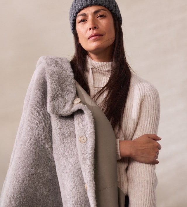 Eine Frau trägt einen hellgrauen Mantel mit weichem Fellkragen über einem cremefarbenen Strickpullover. Das Outfit ist elegant, winterlich und kuschelig-warm.