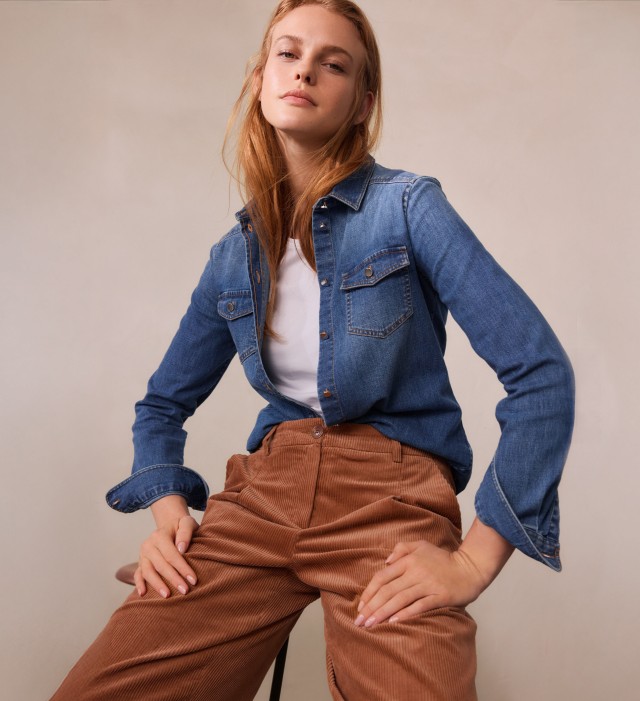 Eine Frau trägt ein blaues Jeanshemd über einem weißen T-Shirt. Dazu kombiniert sie eine braune Cordhose für einen lässigen, retro-inspirierten Look.