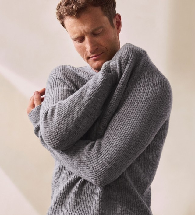 Ein Mann trägt einen grauen, grob gestrickten Pullover mit langen Ärmeln. Das Outfit wirkt warm und gemütlich.