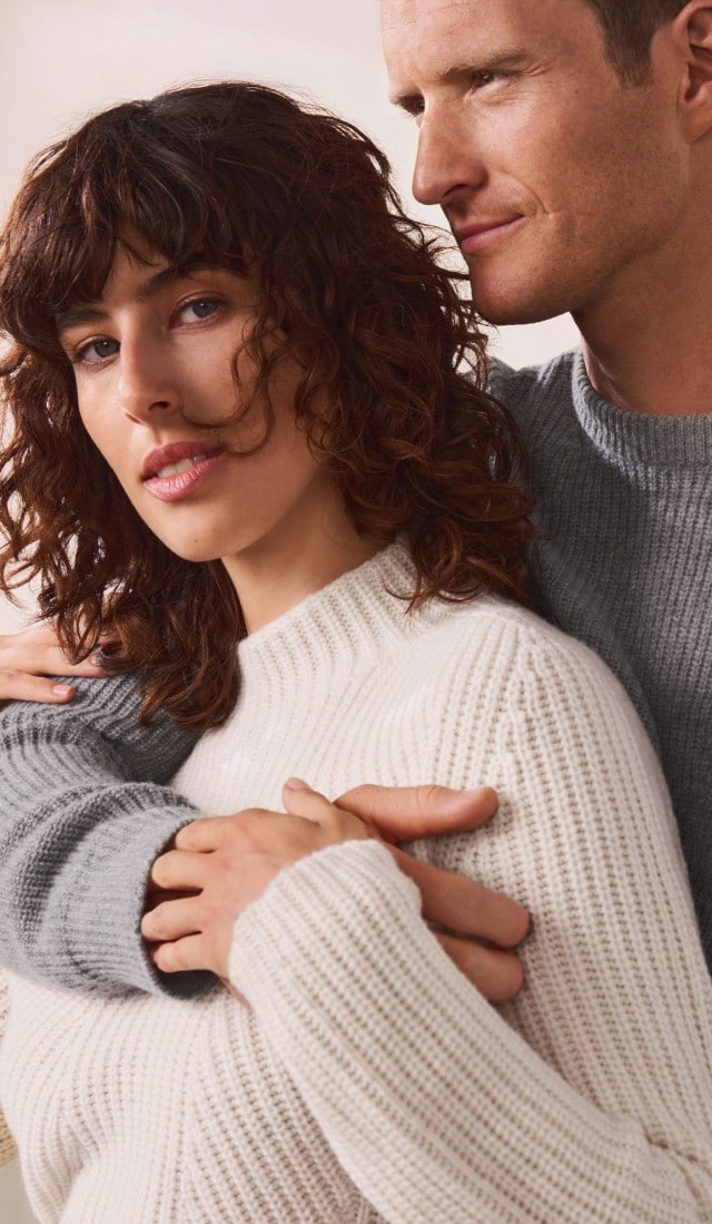Eine Frau trägt einen weißen, grob gestrickten Pullover mit hohem Kragen. Neben ihr trägt ein Mann einen grauen Strickpullover in ähnlichem Stil.