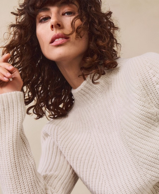 Eine Frau trägt einen weißen, grob gestrickten Pullover mit Rundhalsausschnitt. Der Stil wirkt schlicht und gleichzeitig gemütlich.